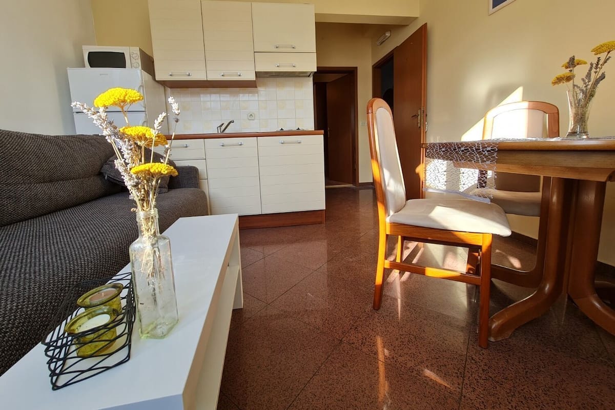 A living room with a sofa, a dining table with a chair, a vase with flowers, and a kitchen in the background.
