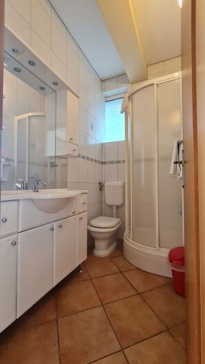 A bathroom with a white sink, mirror, and toilet, a shower with a glass door, and a red trash bin. - Apartments Dami (Orebic)