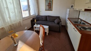En kompakt opholdsstue med sofa, spisebord med blomster, et køkkenområde med vask og komfur samt et vindue med gardiner.