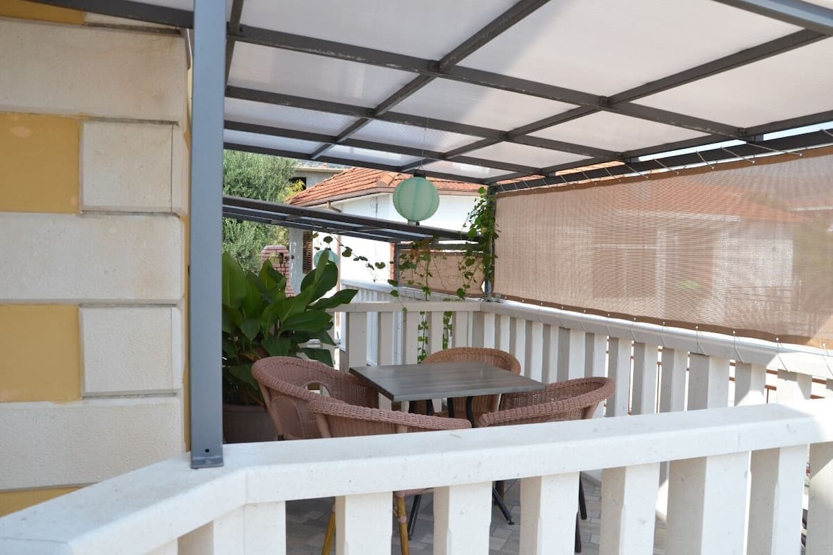 A covered patio with a table and wicker chairs, white railings, and a view of a building with a red-tiled roof.