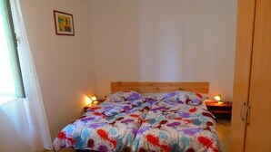 A bed with a colorful bedspread, a wooden headboard, bedside tables with lamps, and a door to the right. - House Kuca za Odmor (Preko)