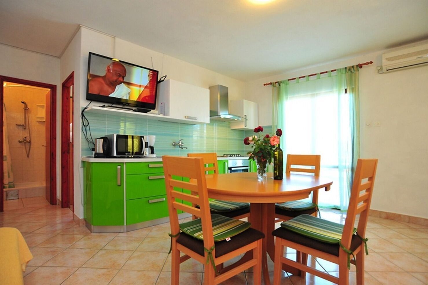Una cocina compacta con armarios verdes, una mesa de comedor con cuatro sillas, un televisor montado sobre la encimera y una ventana con cortinas.