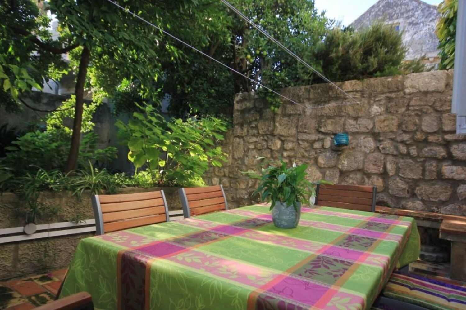 Une terrasse avec une table recouverte d’une nappe colorée, entourée de chaises en bois, et une plante en pot.
