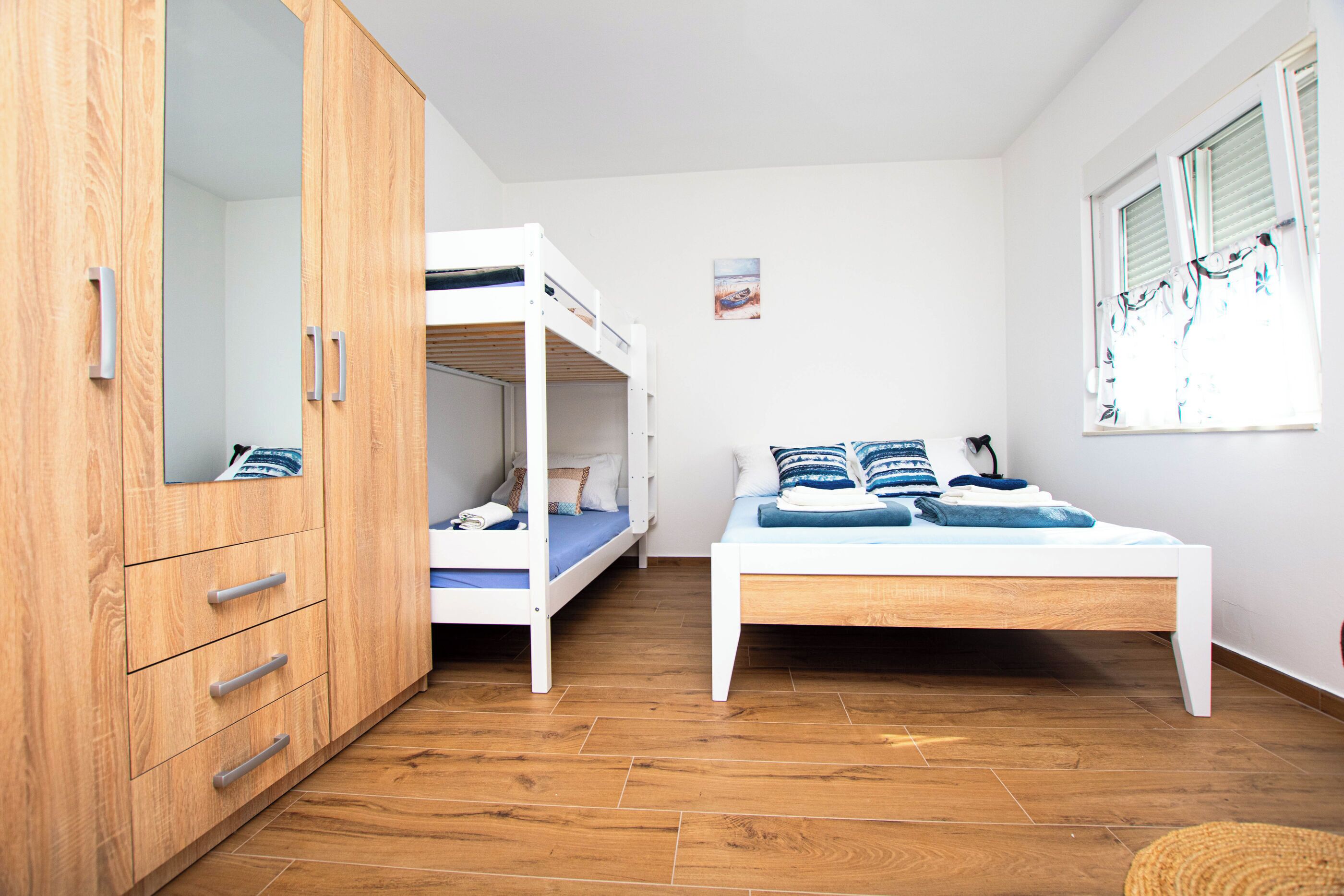 A room with a bunk bed, a single bed, wooden flooring, a wardrobe, and a window with blinds.