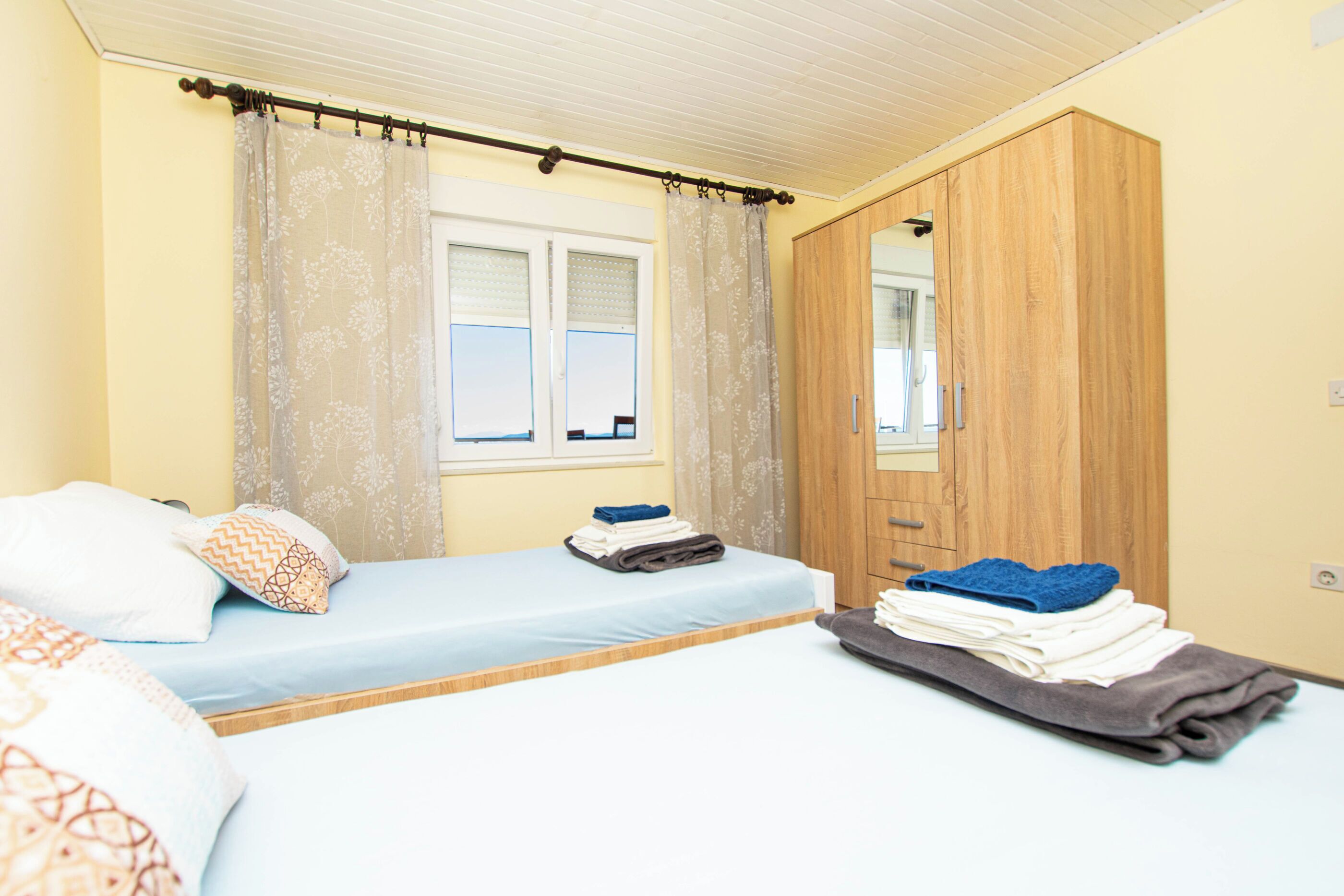 A bedroom with two beds, a wardrobe, a window with curtains, and a mirror.