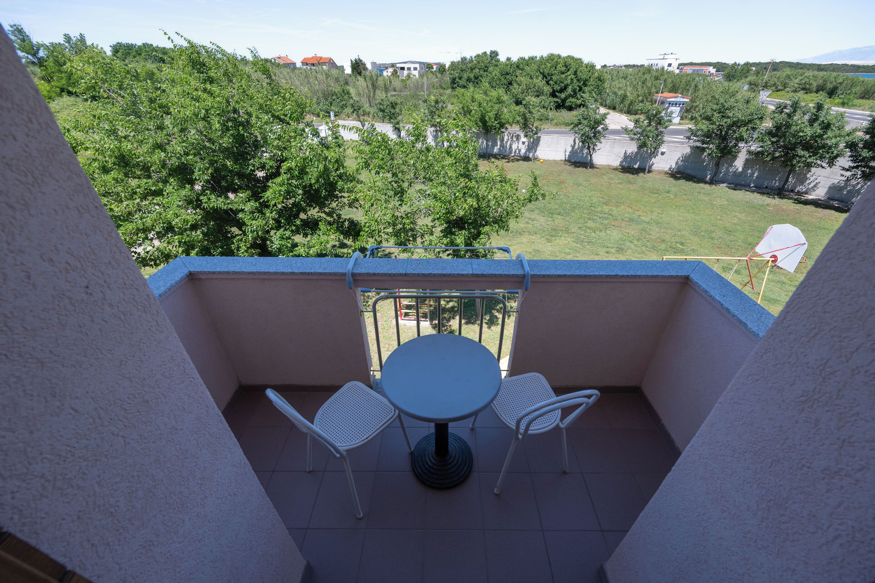 Un balcon avec une table ronde et deux chaises, donnant sur un espace verdoyant agrémenté d’arbres et de bâtiments.