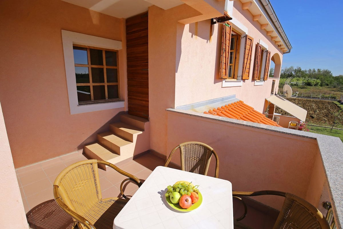 Un balcón con sillas de mimbre, una mesa con una bandeja de frutas y vistas a una casa con techo de tejas.