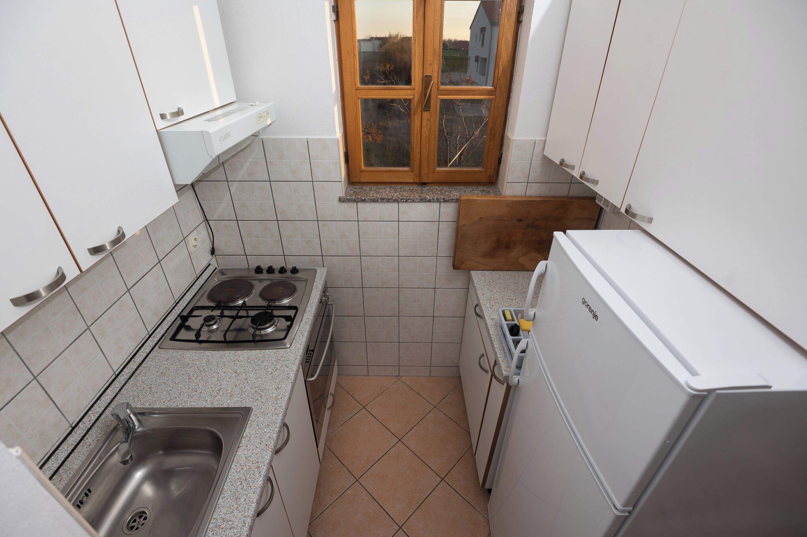Cocina compacta con estufa a gas, lavamanos, refrigerador blanco y ventana con vistas al exterior.