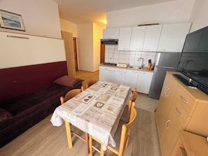A compact living space with a kitchenette, a dining table with a patterned tablecloth, a sofa, and a television. - Apartments Armitage (Privlaka)