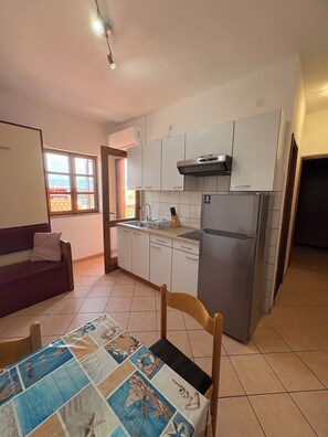 A compact kitchen with a refrigerator, stove, and microwave. There is a window with a view of buildings outside. - Apartments Armitage (Privlaka)
