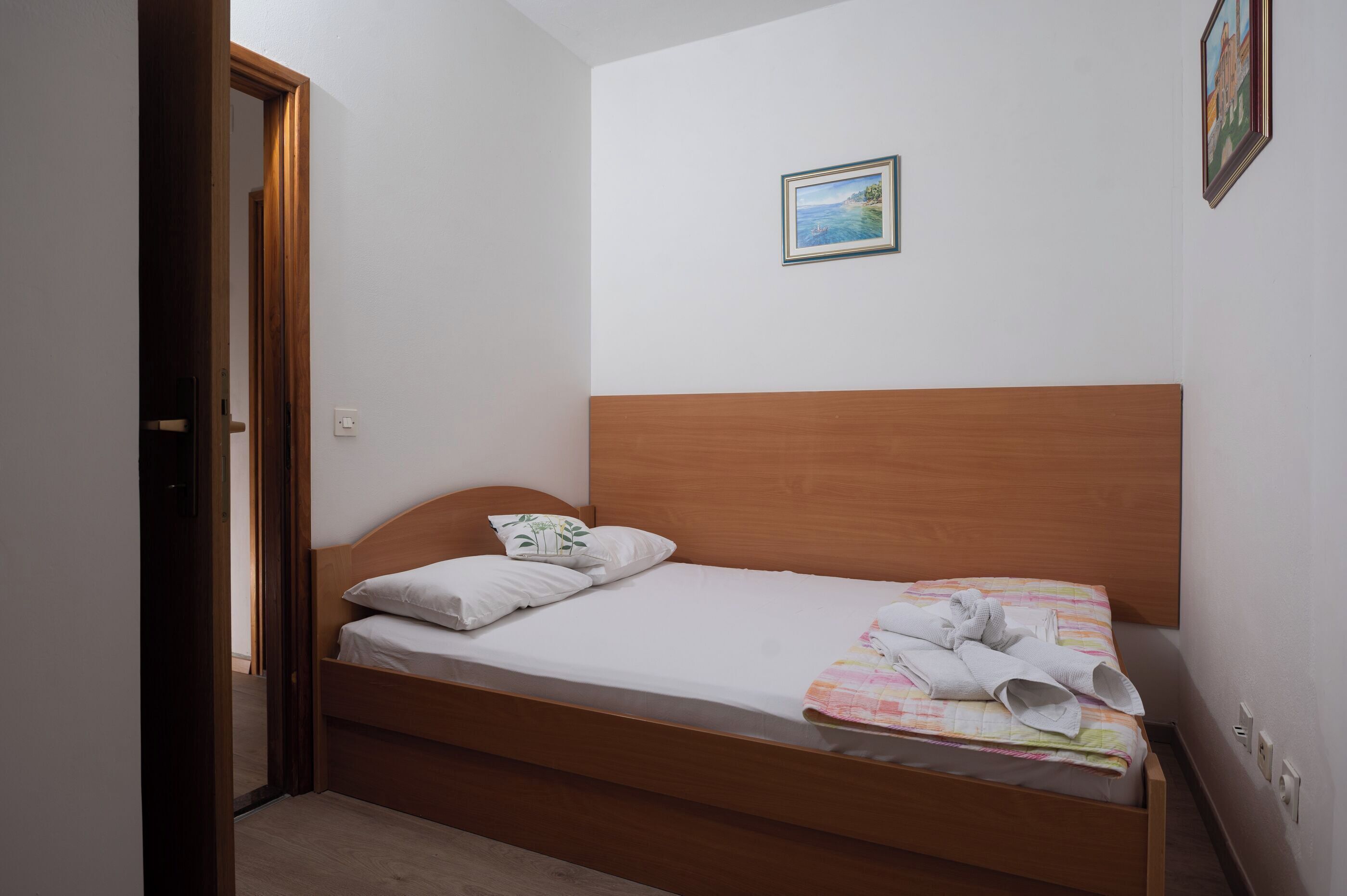 A single bed with a wooden headboard, a small painting on the wall, and a door leading to another room.