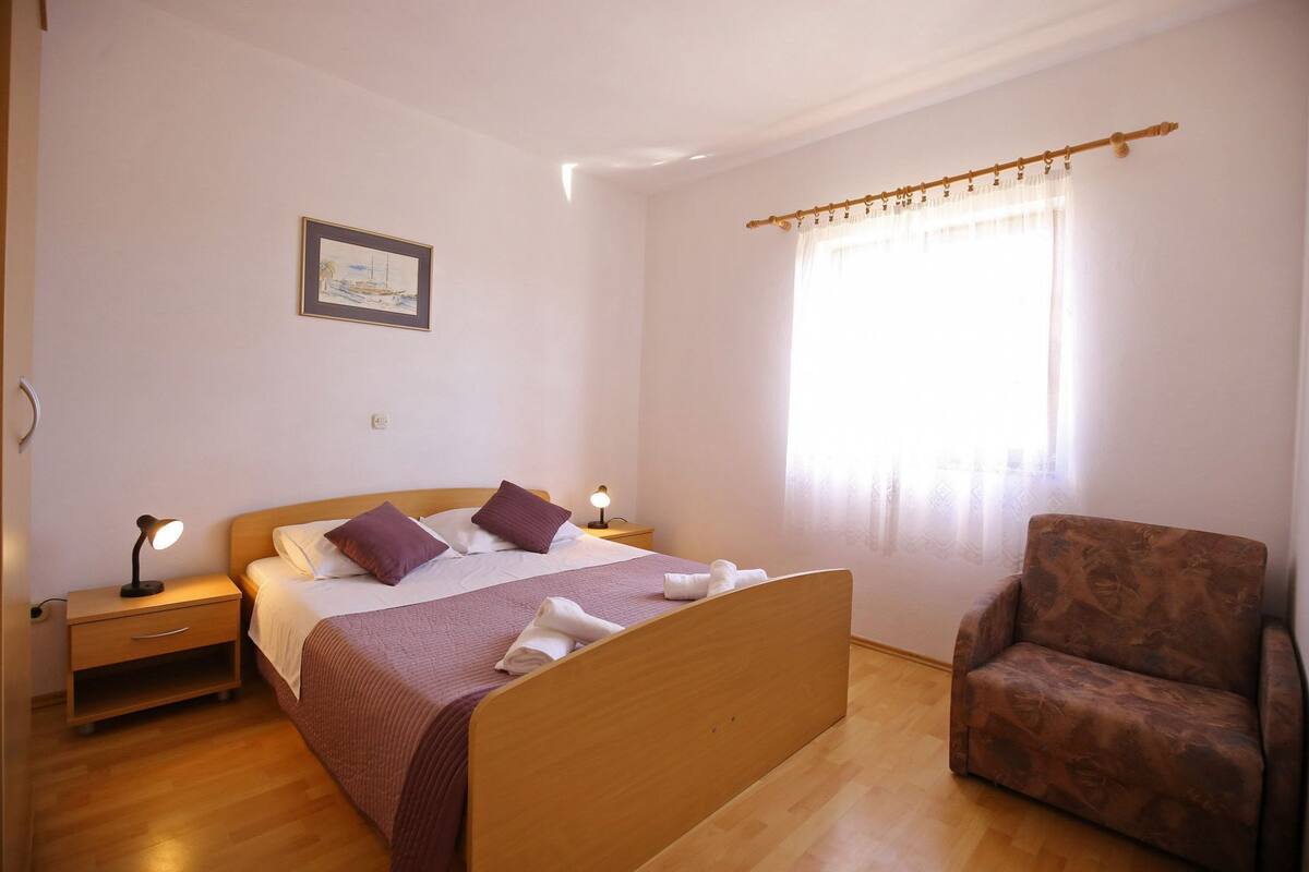 Un dormitorio con cama de madera, dos lámparas de noche, una silla, un ventanal con cortinas translúcidas y un cuadro en la pared.