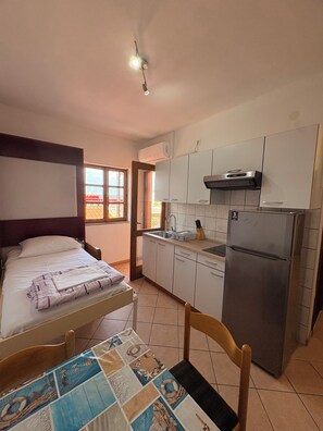 A compact kitchen with a bed, a dining table with a placemat, and a refrigerator. - Apartments Armitage (Privlaka)