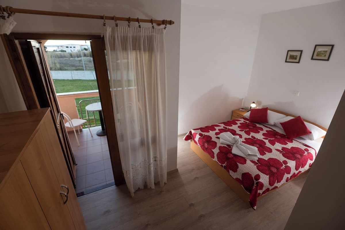 Ein Schlafzimmer mit Bett, Nachttisch, Fenster mit Vorhängen und Blick ins Freie.
