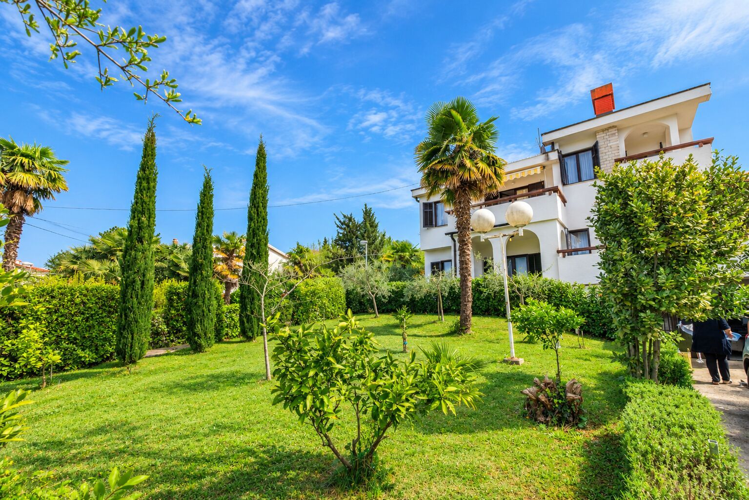 Un jardin bien entretenu, agrémenté de divers arbres et arbustes, un ciel d’un bleu éclatant et un bâtiment à deux étages doté d’un balcon.