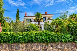 Um edifício de dois andares com um muro de pedra em primeiro plano, uma placa de pare e vegetação exuberante.
