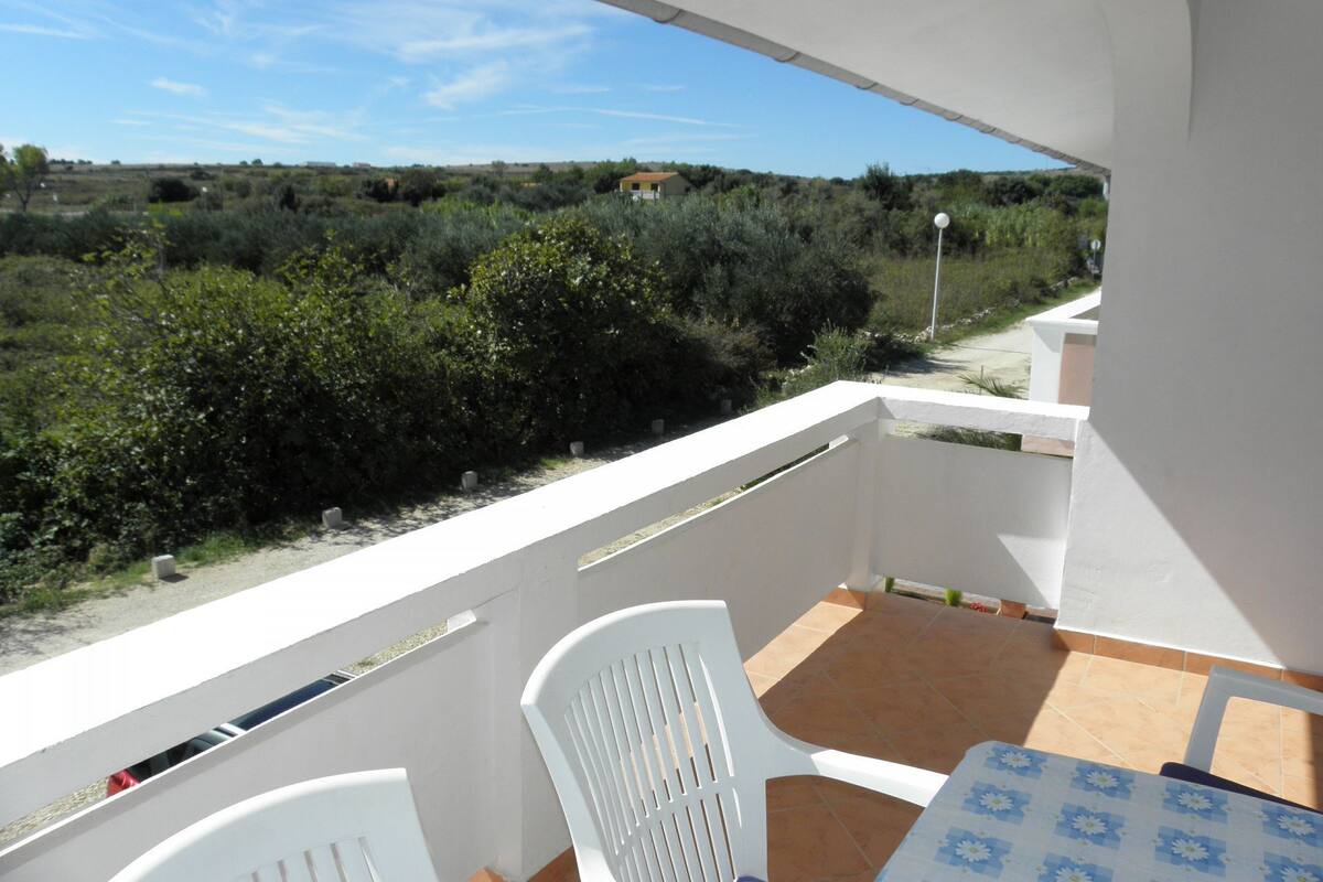 A balcony with a table and chairs, overlooking a landscape with bushes and a distant building.