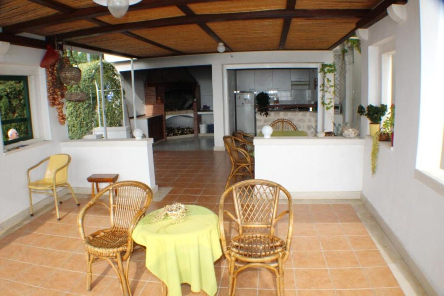 Une terrasse extérieure avec des meubles en osier et une table, un coin cuisine visible à travers une porte ouverte, et des plantes en pot.