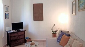 A living room with a sofa, a TV on a stand, a glass table, a lamp, and a painting on the wall. - Apartment Roko (Vis)