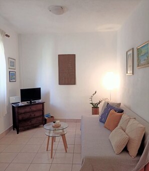 Ein Wohnzimmer mit einer Couch, einem Fernseher auf einem Schrank, einem Glastisch, einer Lampe und einem Bild an der Wand.