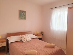 Ein Schlafzimmer mit einem hölzernen Bett, einem Nachttisch, einer Lampe, einem Gemälde und einem Fenster mit durchsichtigen Vorhängen.