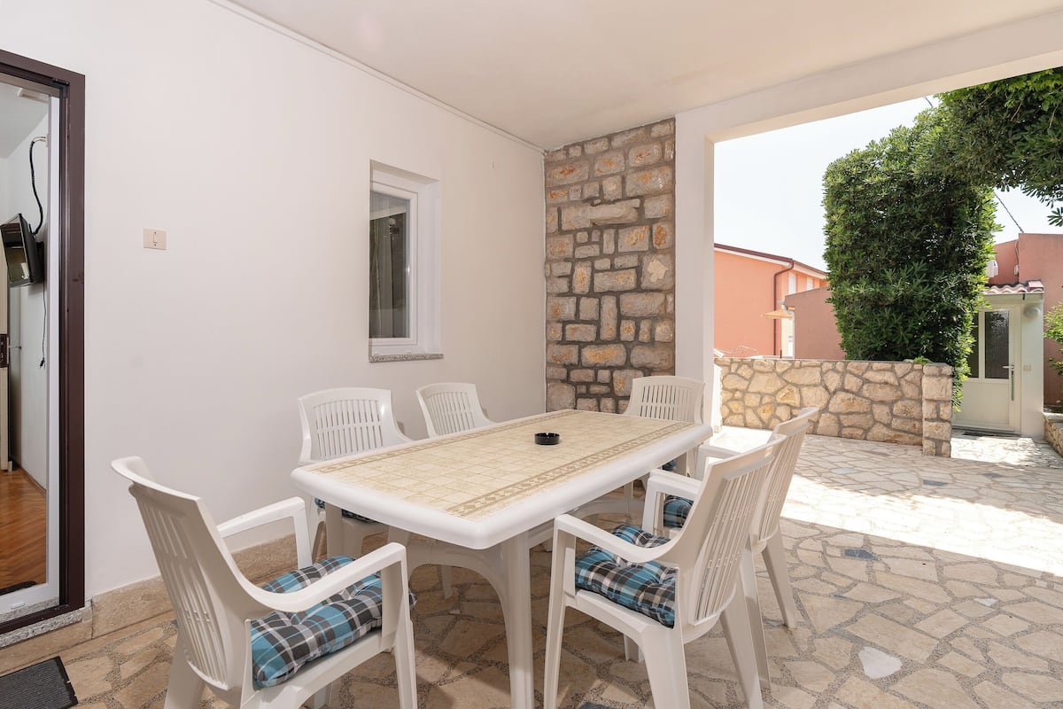 Un coin repas avec une table et des chaises blanches, un mur en pierre et, en arrière-plan, une vue sur un arbre et un bâtiment.