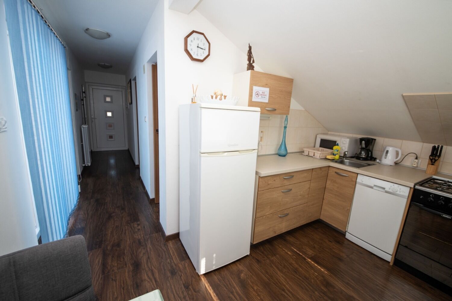 A compact kitchen with a refrigerator, microwave, stove, and dishwasher. There is a clock on the wall and a blue vase on the counter.