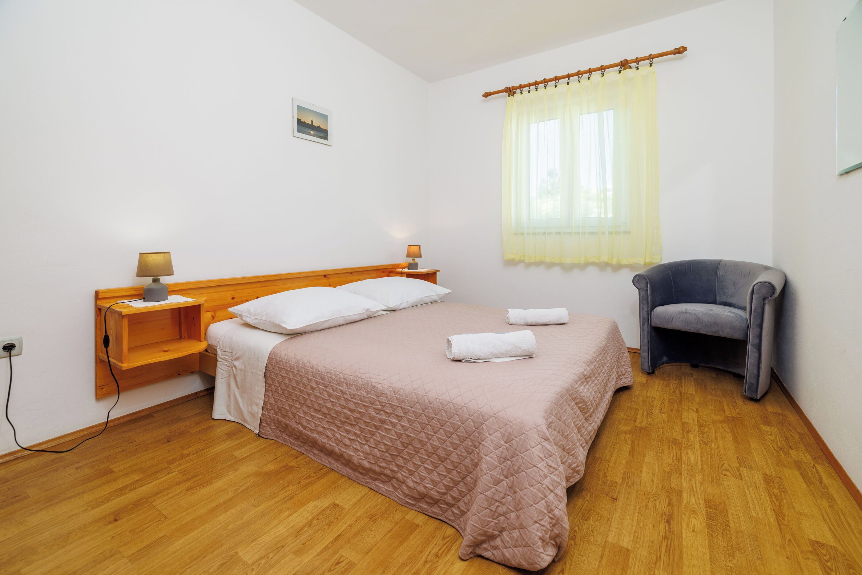 A bedroom with a bed, bedside tables, a chair, a window with curtains, and a wooden floor.