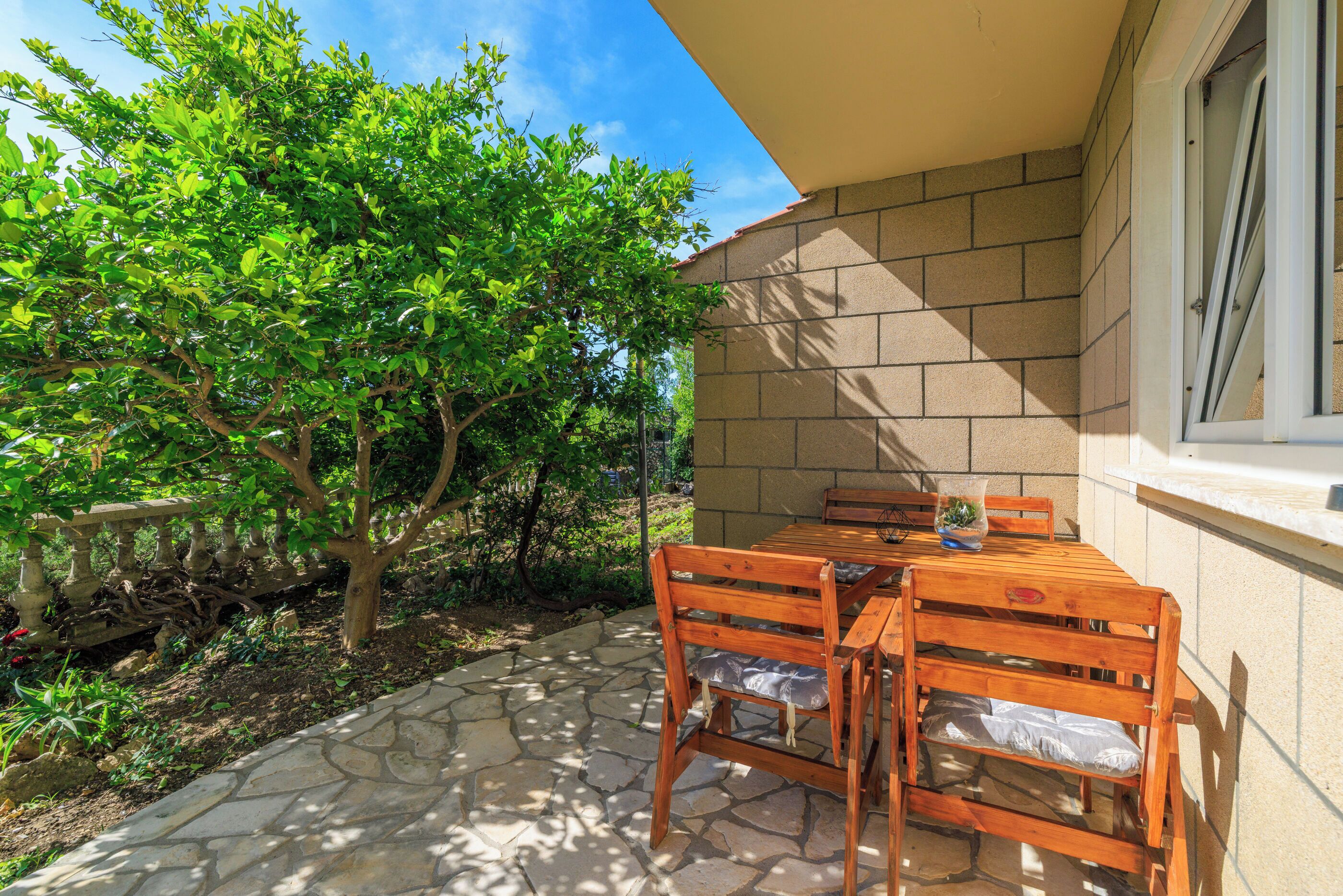 Eine Terrasse mit Steinboden, einem Holztisch und Stühlen sowie einem Baum, der Schatten spendet.
