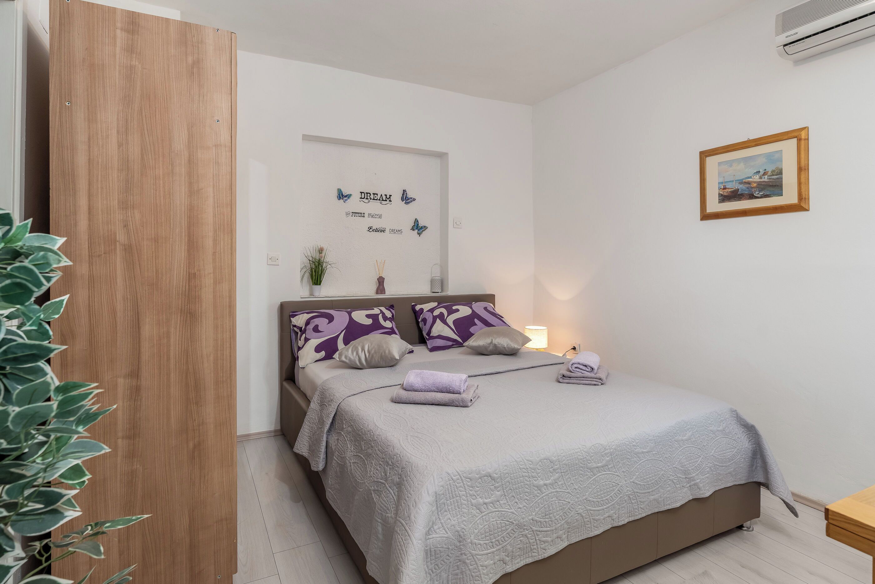 A neatly made bed with a grey quilt, purple patterned pillows, and a small bedside table with a lamp.