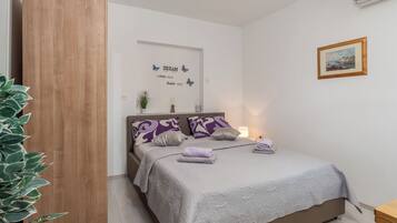 A neatly made bed with a grey quilt, purple patterned pillows, and a small bedside table with a lamp.