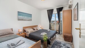 A modern hotel room with a large bed, a smaller bed, a bedside table with a lamp, a vase with flowers, and a view of the sea through a window. - Apartment Don (Pag)