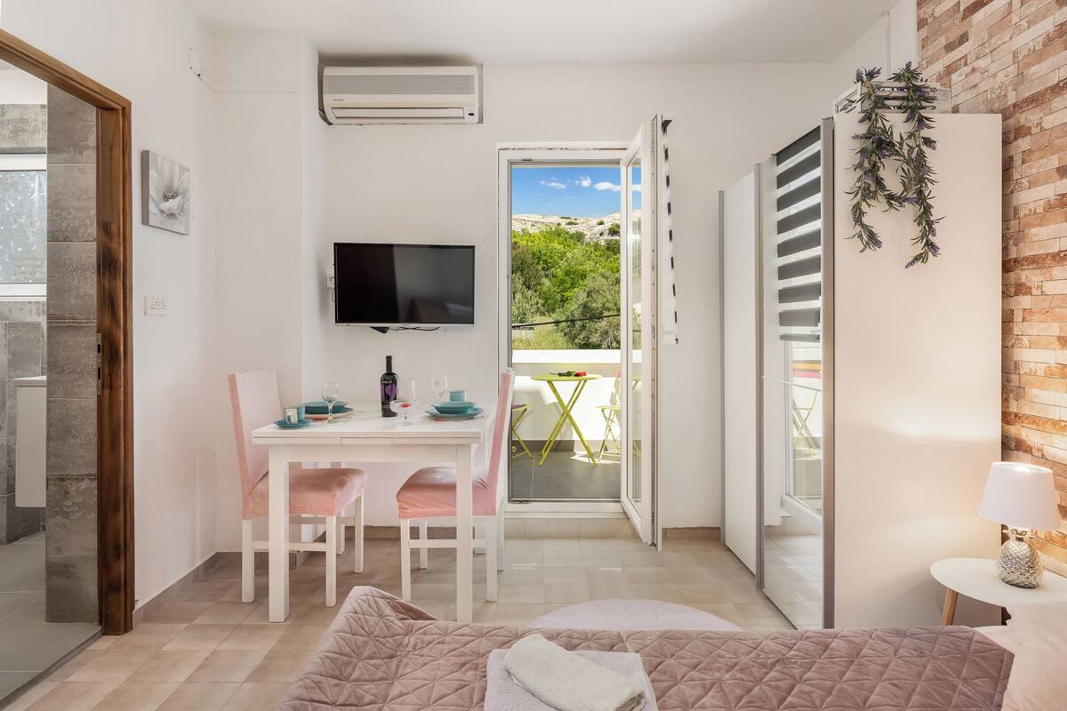 A bedroom with a bed, a desk with a chair, a TV, and a balcony with a table and chairs.