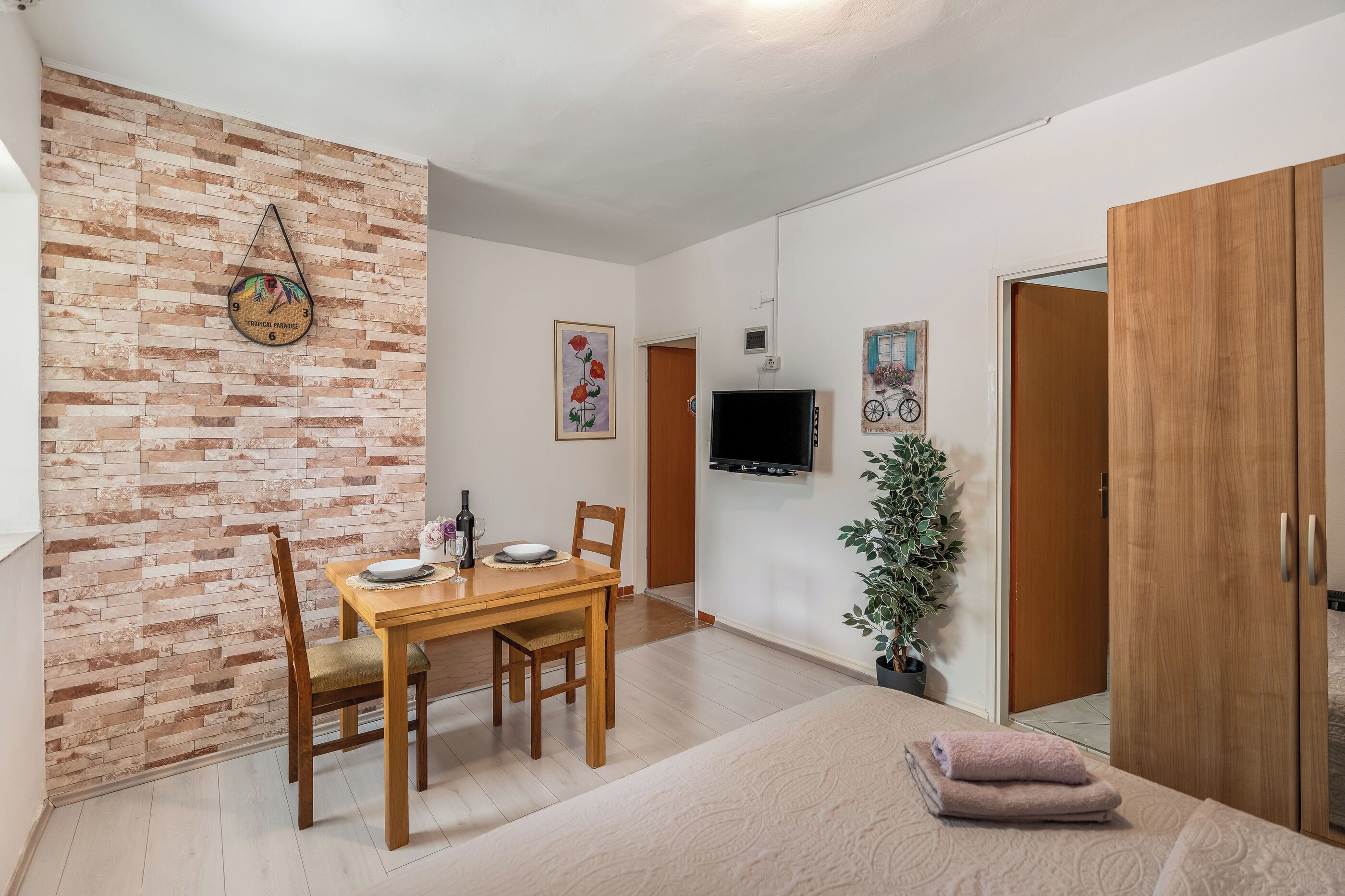 Una habitación con pared de ladrillo, una mesa de comedor con sillas, un televisor de pantalla plana montado en la pared, un armario de madera y una planta en maceta.