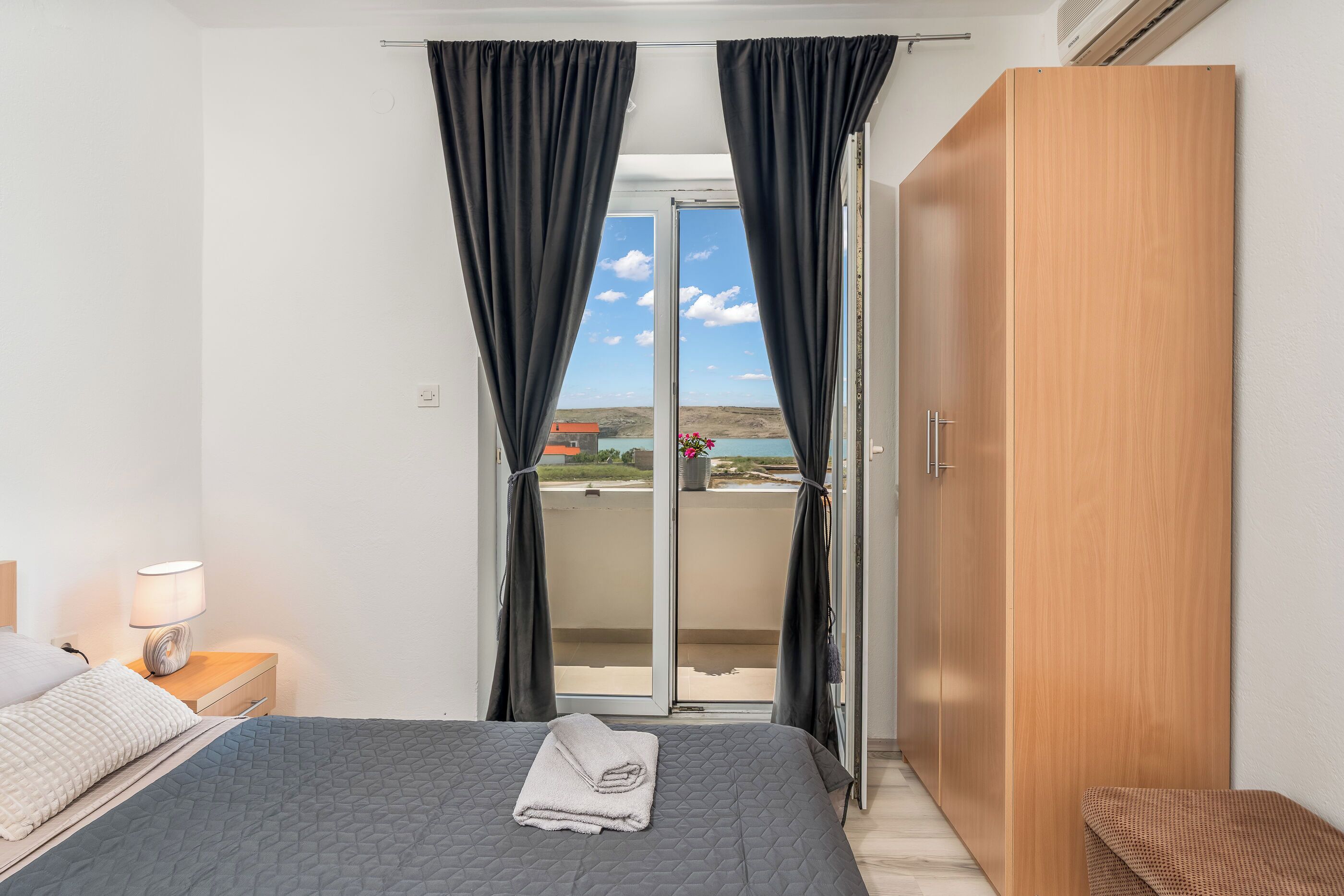 A bedroom with a bed, a nightstand with a lamp, a wooden wardrobe, and a sliding glass door leading to a balcony.