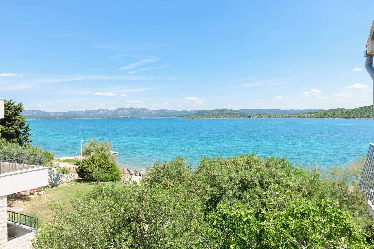A view of a lake with mountains in the background, surrounded by greenery and a building with a balcony.