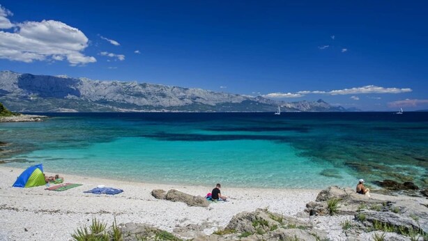 Una spiaggia con acque turchesi cristalline, un litorale roccioso e montagne sullo sfondo.
