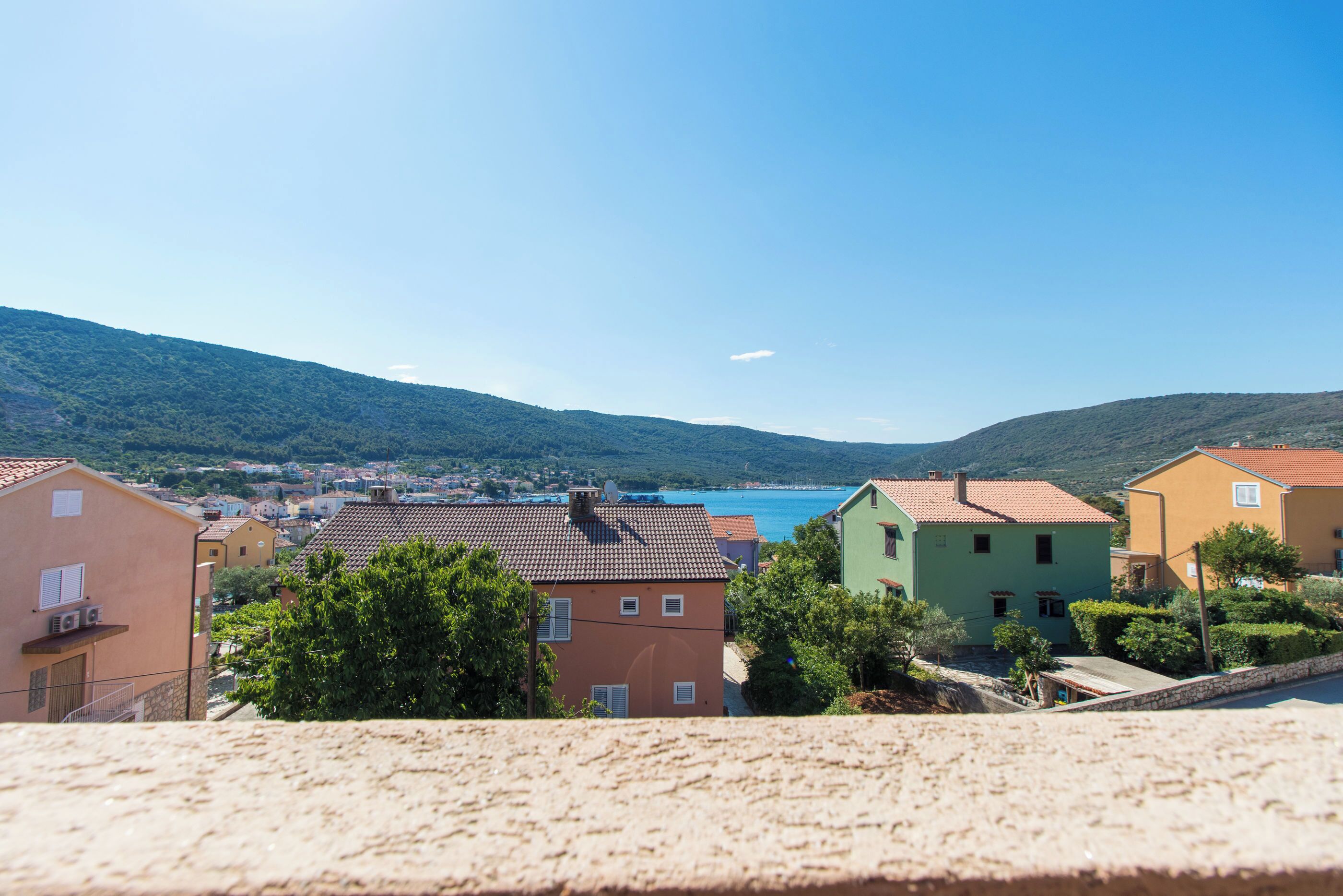 Un poble costaner amb edificis de colors, un cel blau clar i vista al mar i a les colines allunyades.