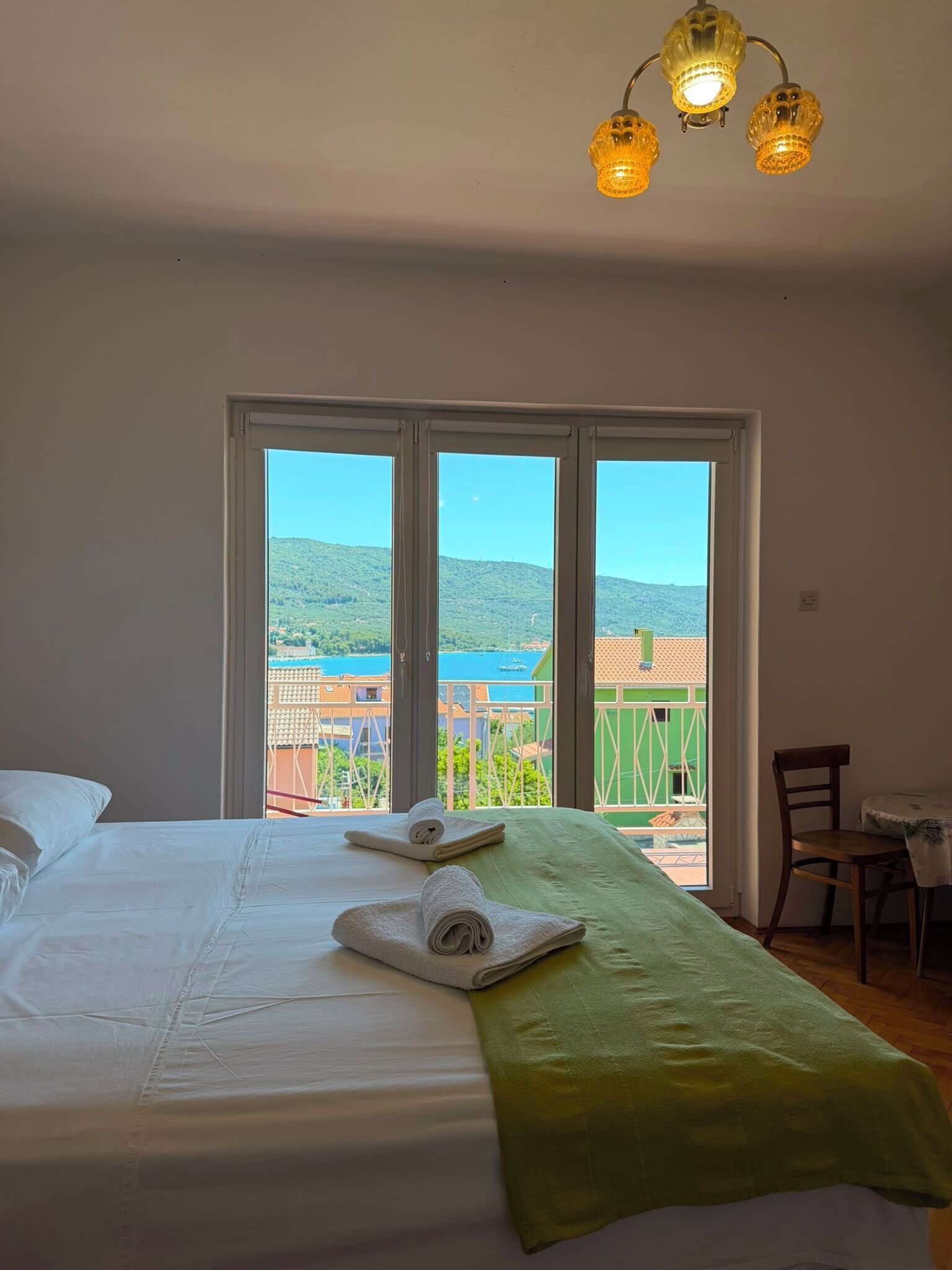 A hotel room with a bed, two towels, a green throw, and a view of a coastal area.