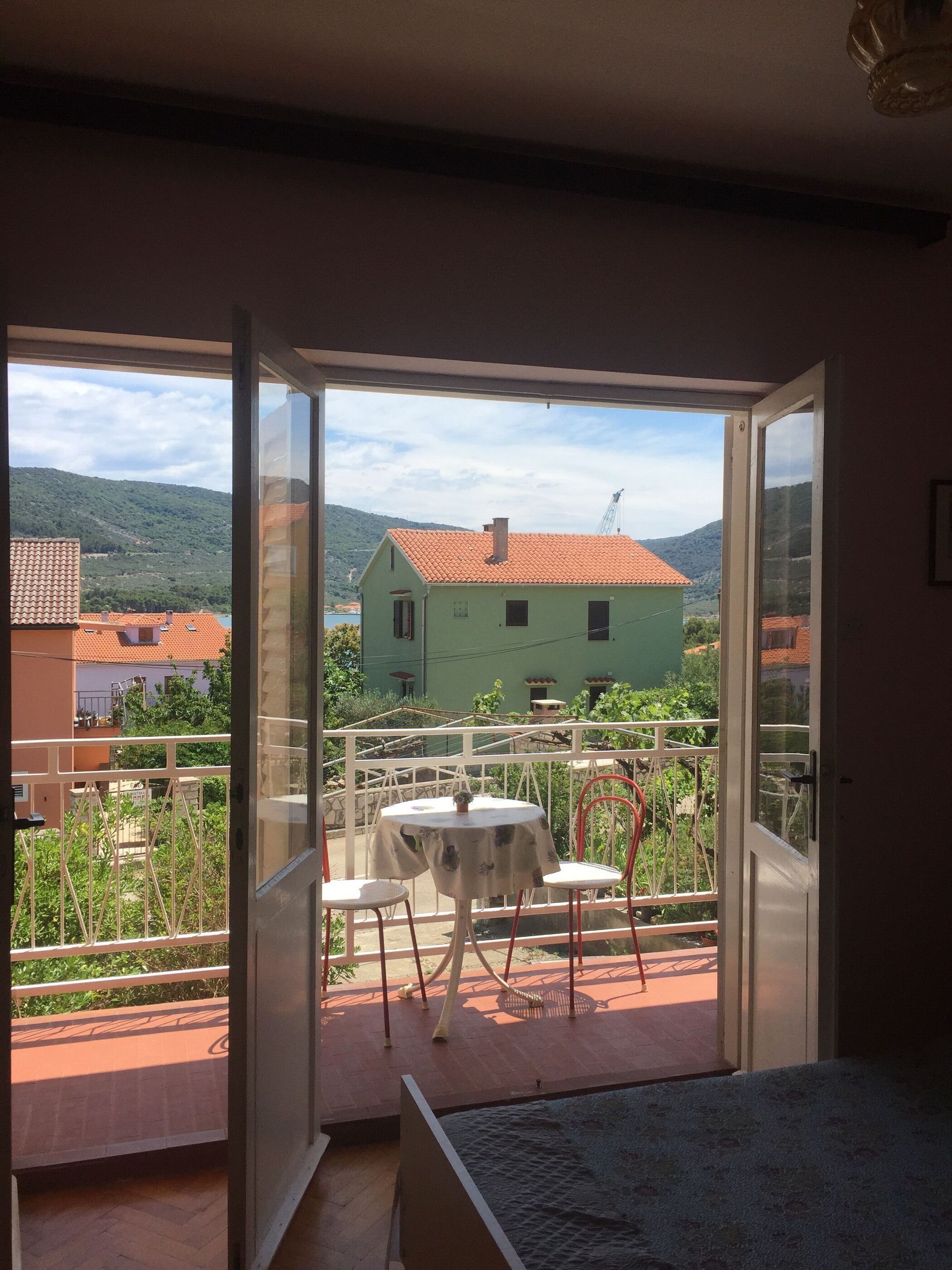 A balcony with a table and chairs, a view of a green house, and a mountainous landscape.