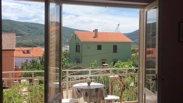 A balcony with a table and chairs, a view of a green house, and a mountainous landscape.