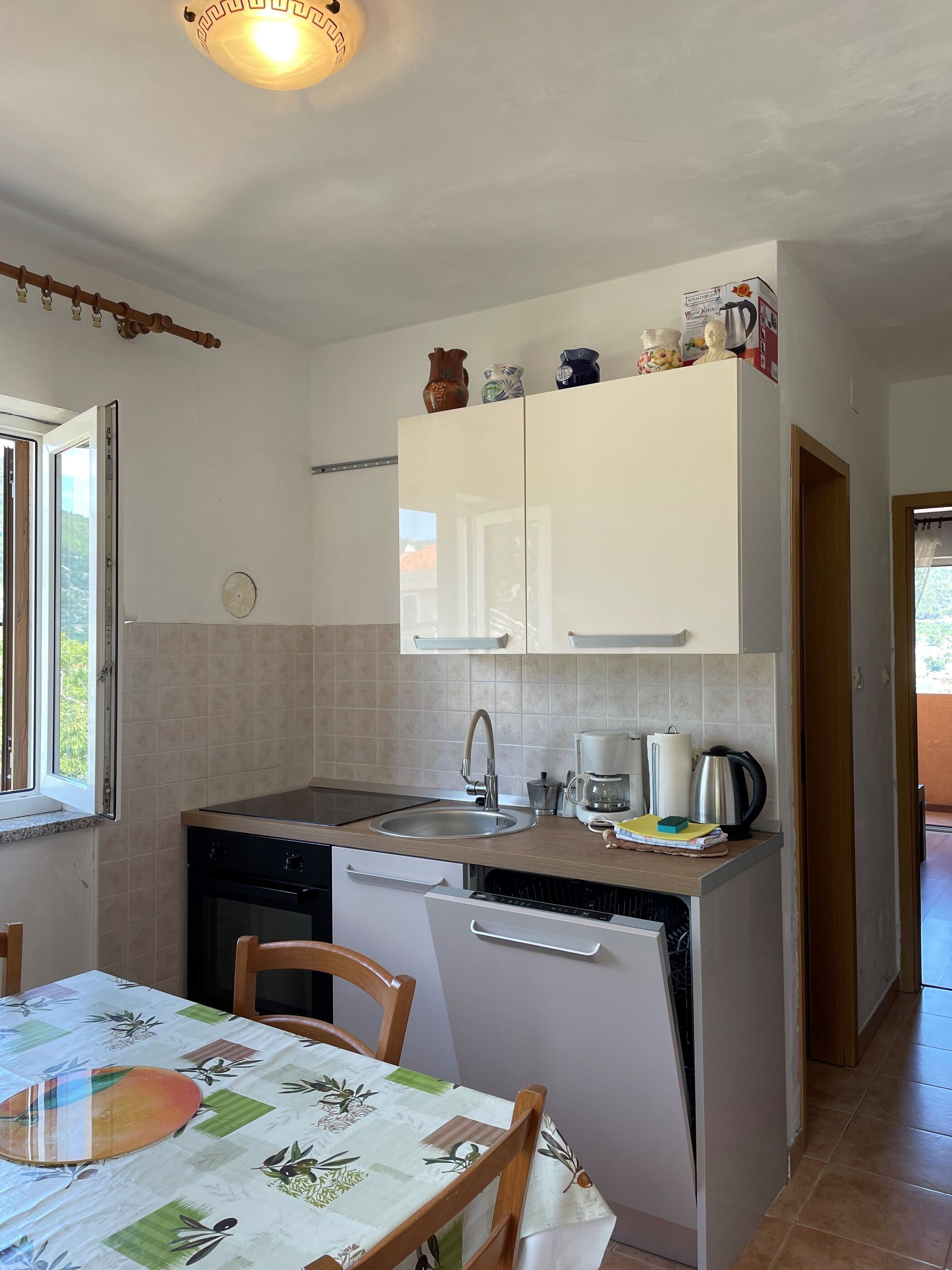 A compact kitchen with a sink, stove, and dishwasher, a dining table with chairs, and a window with a view of greenery.