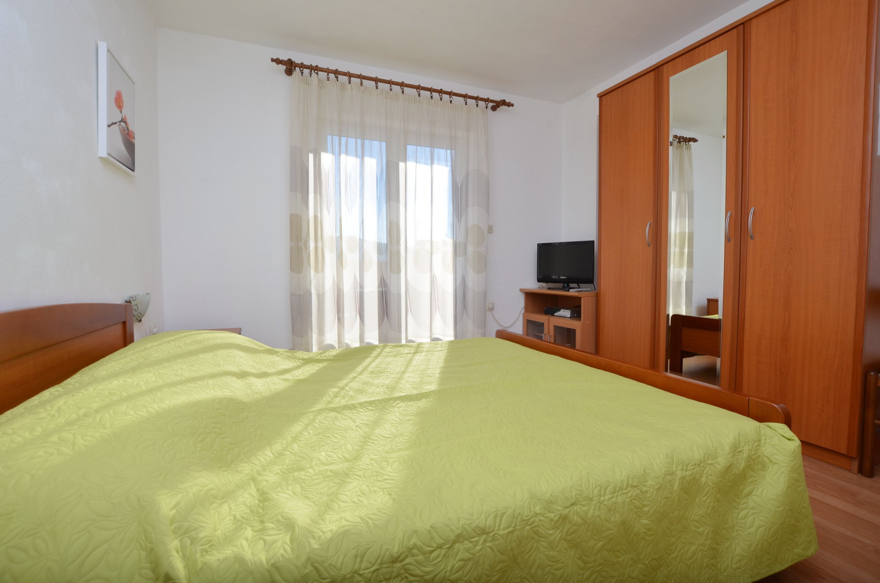 Un dormitorio con cama, televisor, armario y ventana con cortinas.
