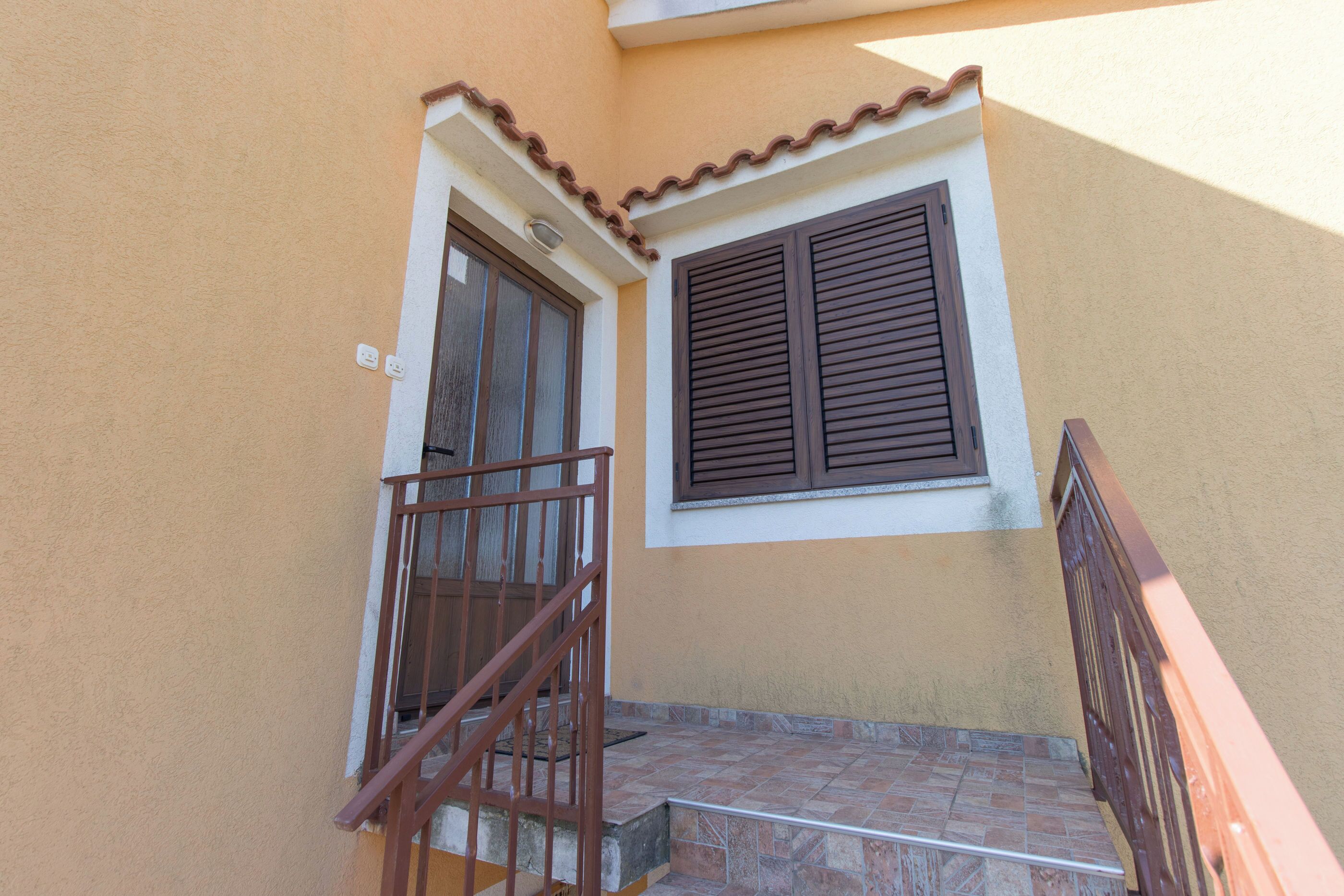 Una escalera con barandilla metálica que conduce a una puerta con un ventanal y contraventanas marrones.