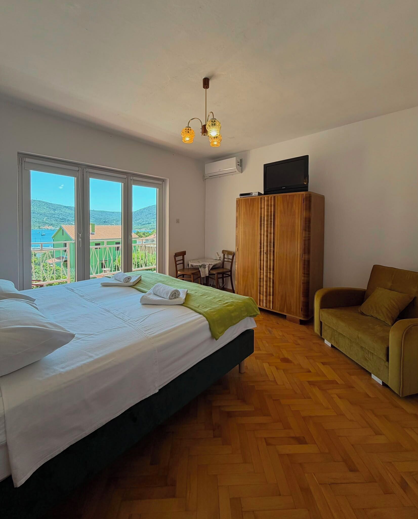 Quarto com cama, guarda-roupa de madeira, televisão, mesa de jantar com cadeiras e vista para o mar através de grandes janelas.
