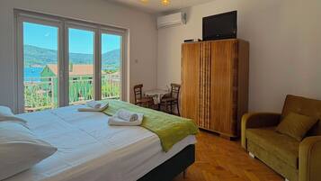Quarto com cama, guarda-roupa de madeira, televisão, mesa de jantar com cadeiras e vista para o mar através de grandes janelas.