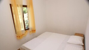 A small, single-bed room with a plain white bedspread and a wooden headboard. A window with sheer curtains is visible, allowing natural light to enter. - Apartments Mari (Blato)