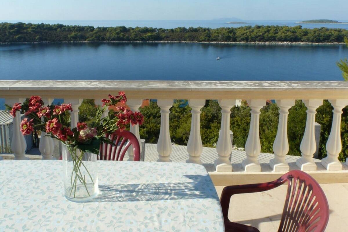 A balcony with a table and chairs, overlooking a calm body of water and a forested coastline.