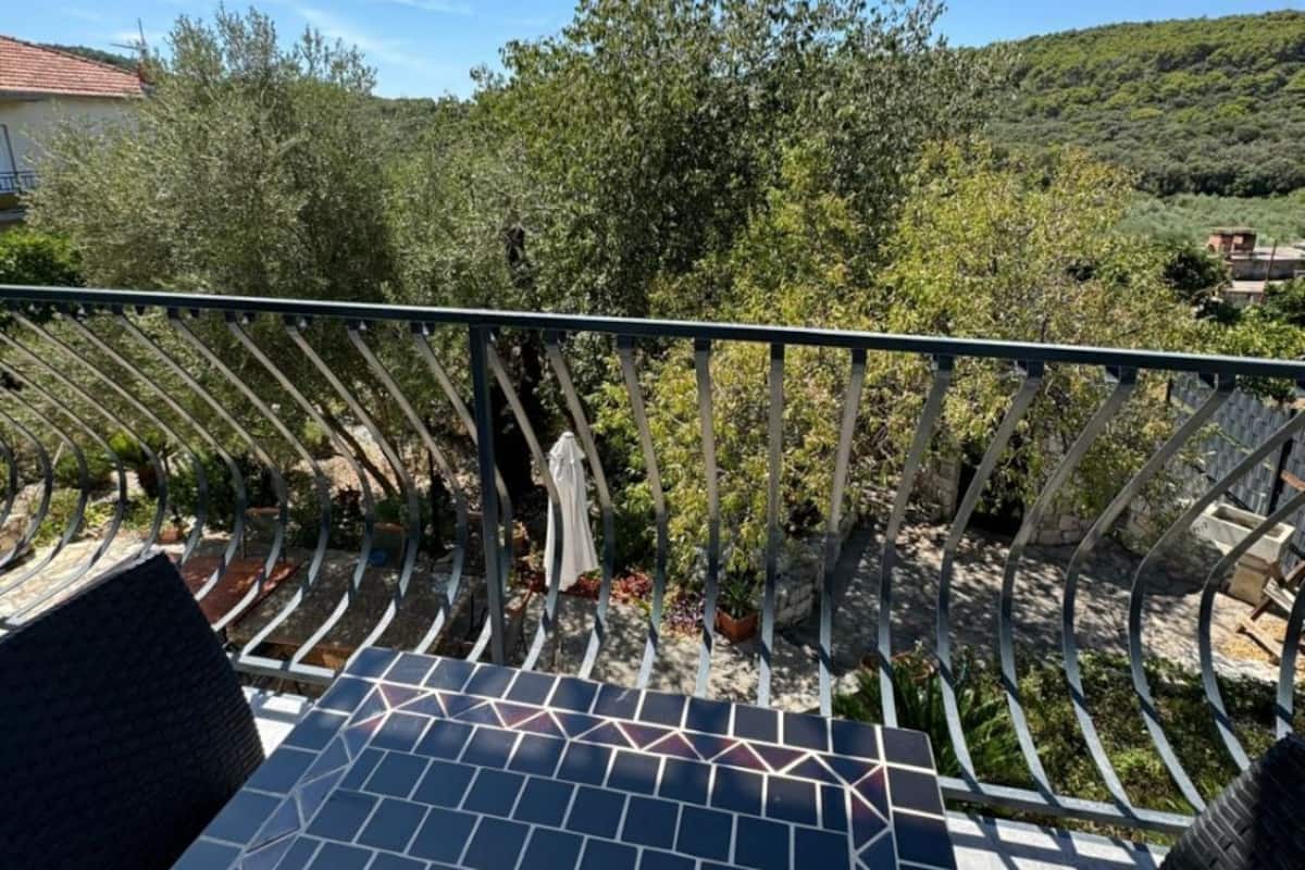 Un balcón con piso de baldosas, baranda de metal y vistas a una colina con árboles y edificios.