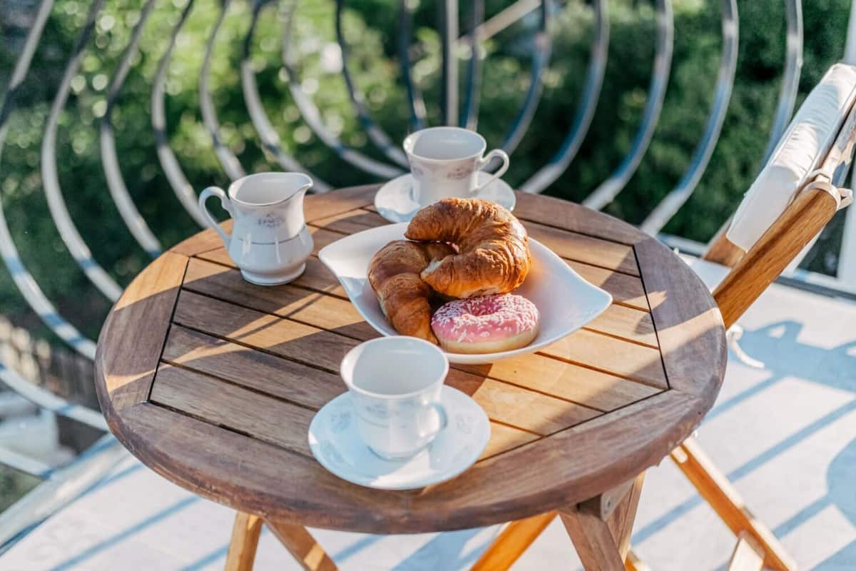 Una mesa redonda de madera con un plato blanco que contiene croissants y una rosquilla, dos tazas blancas y una tetera blanca.